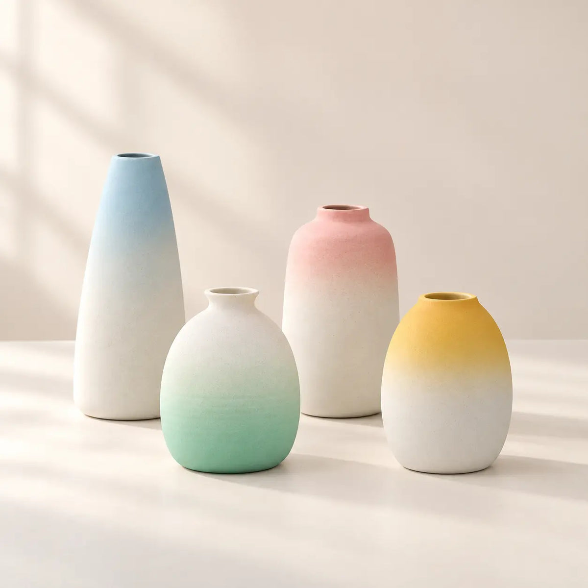 Four pastel gradient ceramic vases in blue, blush pink, mint green and yellow on an ivory surface.