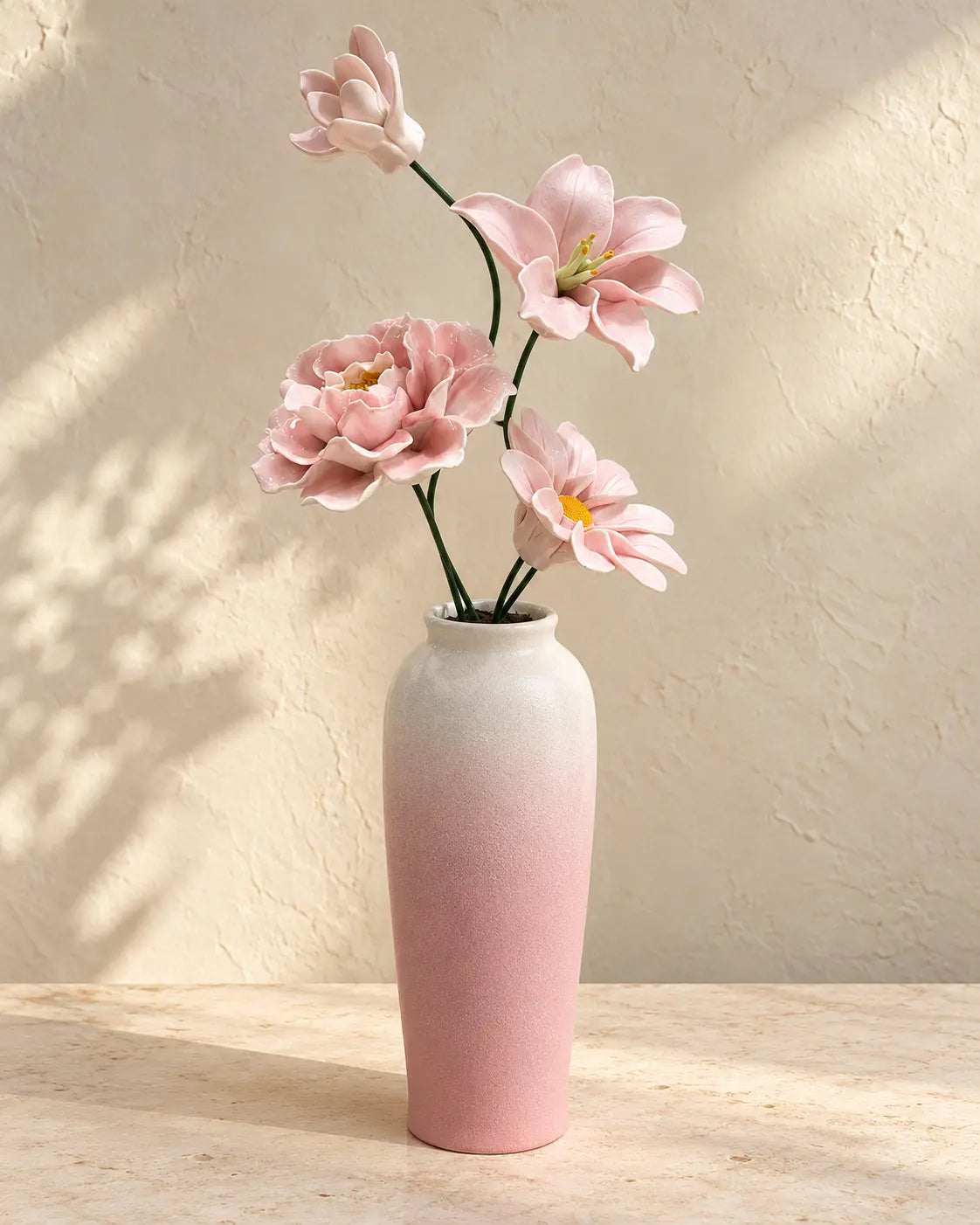 Blush pink porcelain flowers in a tall ivory-to-pink gradient vase on a pale stone tabletop.